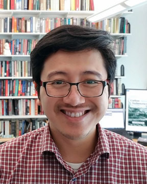 A man wearing glasses and a red checked shirt, with bookshelves and two computer monitors in the background.